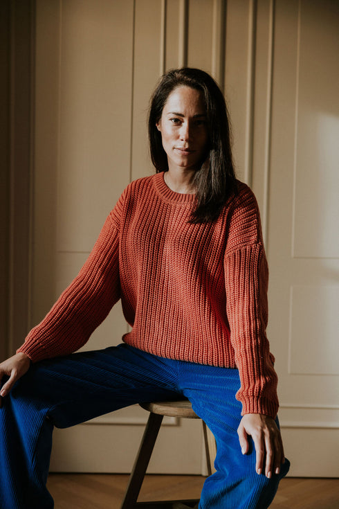 A woman with long dark hair sits on a chair in a softly lit room with light paneled walls, wearing a red knit sweater and Yuki kidswear WOMEN'S Trousers Beau Corduroy in cobalt, looking confidently at the camera.