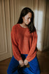 A woman with long dark hair sits indoors, wearing a loose orange sweater and Yuki kidswear WOMEN'S Trousers Beau Corduroy in Cobalt. Calmly gazing down under soft light, she embodies relaxed slow fashion style.