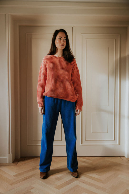 A woman with long brown hair stands indoors on a wooden floor, facing the camera and wearing a loose pink sweater and Yuki kidswear WOMEN'S Trousers Beau Corduroy in cobalt, embodying slow fashion by white double doors.