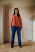 A woman with long brown hair stands indoors on a wooden floor, facing the camera and wearing a loose pink sweater and Yuki kidswear WOMEN'S Trousers Beau Corduroy in cobalt, embodying slow fashion by white double doors.