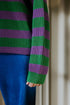 A person wears a green and purple striped ribbed sweater and Yuki kidswear WOMEN'S Trousers Beau Corduroy - COBALT, showing only the lower arm and torso. The partially covered hand highlights a slow fashion style.
