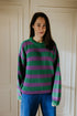 A person with long dark hair stands indoors, smiling gently in front of light-paneled doors, wearing a purple and green striped sweater and Yuki kidswear's WOMEN'S Trousers Beau Corduroy in COBALT.