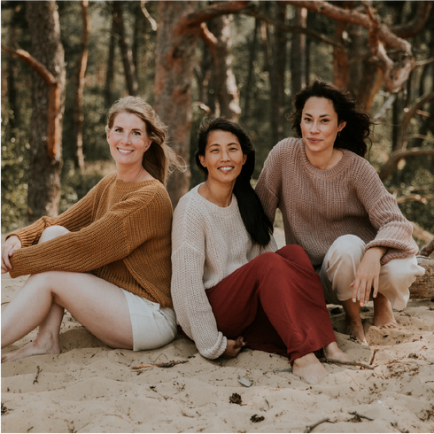 Three women sit barefoot in a wooded area, smiling at the camera. They wear cozy, slow fashion pieces like the Chunky Knitted WOMEN'S Sweater - MOON from Yuki kidswear, enjoying a peaceful outdoor moment among the trees.