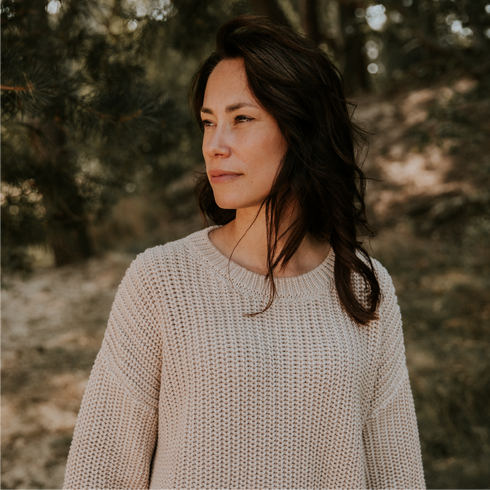 A woman with wavy brown hair wears the Chunky Knitted WOMEN'S Sweater - MOON by Yuki kidswear, standing outdoors amid sunlit trees and gazing thoughtfully to the side in a serene, natural setting.