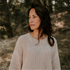 A woman with wavy brown hair wears the Chunky Knitted WOMEN'S Sweater - MOON by Yuki kidswear, standing outdoors amid sunlit trees and gazing thoughtfully to the side in a serene, natural setting.