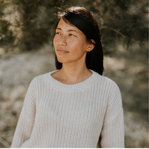 A woman with long dark hair wears the Yuki kidswear Chunky Knitted WOMEN'S Sweater - MOON while standing outdoors, sunlight filtering through trees in a soft, natural setting as she gazes thoughtfully into the distance.